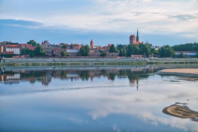 Peaceful reflection of Torun Old Town on Wisla River under a serene summer sky.